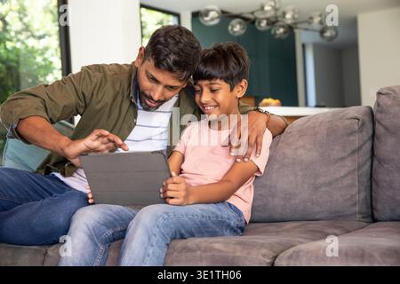 Asian father and son sitting on grey sectional sofa using tablet in home living room. Family, bonding, relaxation, contemporary, lifestyle, comfort, digital || Model released Stock Photo