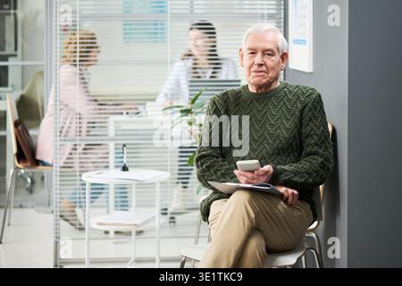 Senior Man Sitting in Waiting Room Using Smartphone and Holding Clipboard Stock Photo