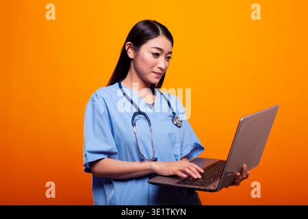 Female nurse wearing blue scrubs and using a laptop against a yellow background. Young asian woman with stethoscope around her neck, standing and typing on her personal computer. Stock Photo