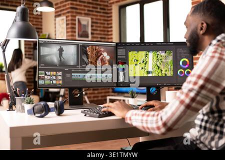 Black male vfx artist works remotely at desk, mixing and adding visual effects to video clips. Creative african american editor using dual monitors for efficient post production workflow. Stock Photo
