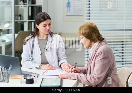 Senior Woman Signing Medical Consent Form with Female Doctor Present Stock Photo