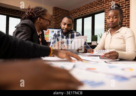 Black professionals collaborate at table, reviewing and analyzing financial charts and reports during business evaluation meeting in modern workspace. Focused team members work together in office. Stock Photo