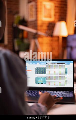 African American woman works remotely from cozy apartment in evening, reviewing company stock market listings on laptop. Female entrepreneur checks business data and online statistics. Stock Photo