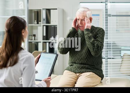 Senior Man Touching Head While Consulting Female Doctor in Clinic Stock Photo