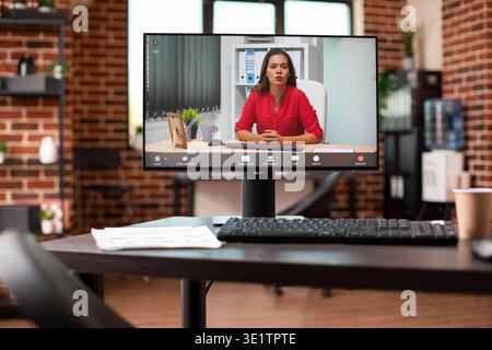 Videoconference call with businesswoman is seen on desktop pc in vacant brick wall office. Closeup on computer screen displaying virtual meeting of female entrepreneur, doing a business presentation. Stock Photo