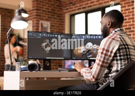 Creative male developer using dual monitors, reviewing gears and equipment while designing construction prototypes. African American engineer visualizing 3D model at home workstation. Stock Photo