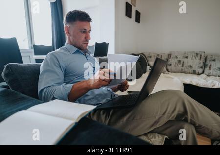 Man working from home on couch with laptop and documents, reviewing paperwork and planning Stock Photo