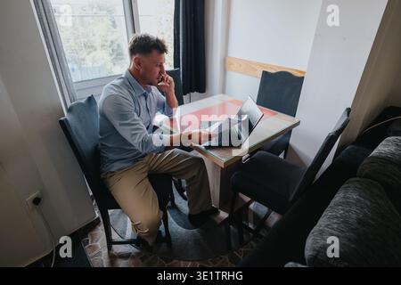 Man working from home on laptop and phone at a small dining table handling documents Stock Photo