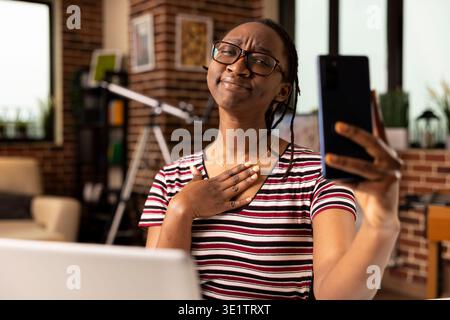 African american female manager places hand on chest with thankful expression while speaking to colleagues through cellphone. Remote freelancer showing appreciation during virtual meeting. Stock Photo