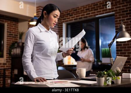Focused employee reading a document in hand, standing at her office desk during her night shift. Asian corporate manager on her feet, working late, planning a brand strategy for startup company. Stock Photo