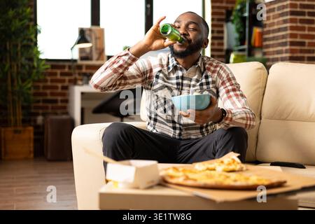 Black person drinking beer with bowl of chips in hand, enjoying relaxing weekend at home. African american man sitting comfortably on sofa having a beverage and snacks in brick wall apartment. Stock Photo