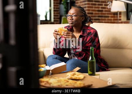 Smiling black woman relaxing on couch, grasping burger and and watching tv show with concentration. Young lady spends time at home enjoying cozy entertainment, takeaway meal and peaceful downtime. Stock Photo