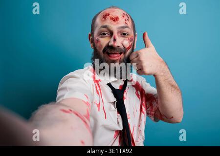 Apocalyptic monster doing thumbs up sign in front of camera, taking picture on smartphone. Creepy evil brain eating corpse showing like and approval symbol, scary horror zombie. Stock Photo