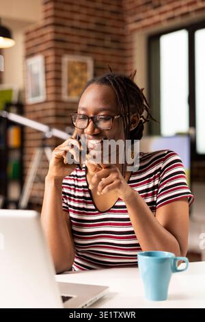 African american woman having cheerful conversation on cellphone while working on laptop in home office. Happy female entrepreneur seated at desk, talking on mobile device with business investor. Stock Photo