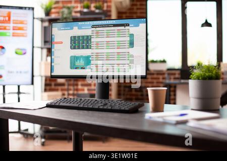 Empty workspace with desktop pc and big digital screen desk displaying business analytics data. Office table with nobody, has computer screen showing company statistics information in brick wall room. Stock Photo