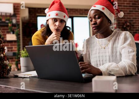 Colleagues wearing santa hats working on laptop for project deadline during christmas holiday. Two diverse women employees in decorated office during festive corporate season Stock Photo