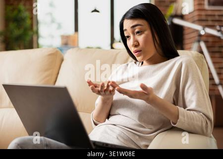 Female financial advisor sitting with laptop in brick wall apartment, engaged in virtual meeting with startup investors. Asian woman manager discussing business strategy remotely from home. Stock Photo