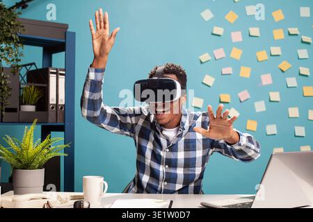 Excited employee uses VR headset, blending technology with workplace creativity. African american man exploring virtual reality, fully engaged in an interactive digital world at his office desk. Stock Photo