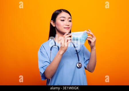 Asian female nurse, wearing blue scrubs and stethoscope, poses confidently with face mask in her hands. Standing against yellow isolated backdrop, she looks ready for her role in healthcare. Stock Photo