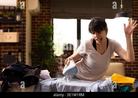 Excited caucasian woman enjoying household chores, dancing to music while ironing pile of clothes. Young female adult filled with positive energy doing laundry and removing wrinkles from shirts. Stock Photo