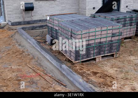 Stacked concrete paving stones secured with green straps on wooden pallets beside a partially constructed pathway at a residential building site Stock Photo