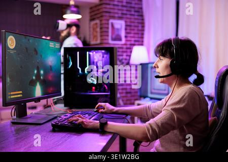 Gamer using professional grade level gaming PC to avoid disruptive lag while playing online esports matches. Woman relaxing in neon lights living room playing action shooting videogame late at night Stock Photo
