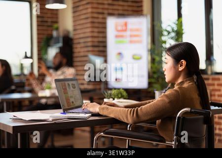 Businesswoman with a disability uses her laptop at desk, analyzing company growth charts and statistics. Asian female entrepreneur in a wheelchair reviewing financial data in brick wall workspace. Stock Photo