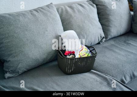 A dark storage basket filled with colorful yarn balls and knitting supplies, placed on a grey fabric sofa with soft cushions. Warm and cozy home atmosphere for DIY projects, handmade crafts, and creative hobbies. Knitting Basket with Yarn Balls on Grey Sofa. High quality photo Stock Photo