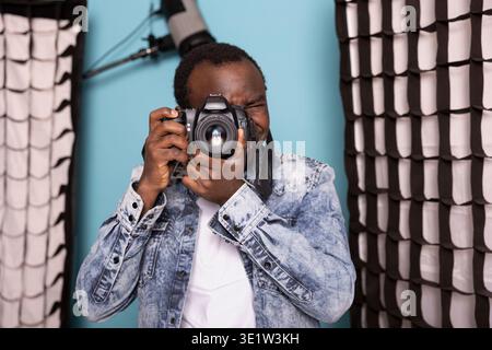 Black male photographer, deeply focused, using his DSLR camera in a studio setting. African american man positioned near softboxes and professional lighting, preparing to capture high quality images. Stock Photo