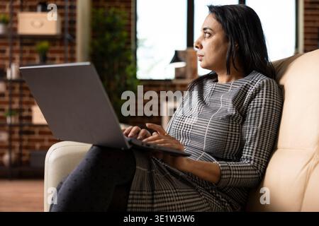 South asian businesswoman sits on couch with laptop, appearing distracted, looks around brick wall apartment. Remote female employee loses focus while working from home in cozy living room. Stock Photo