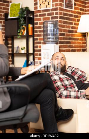 White man lies on couch in private clinic, attentively listening to psychologist during therapy session. Caucasian male patient receives guidance and mental health evaluation from therapist. Stock Photo