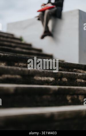 Person sitting on concrete ledge above weathered steps holding a red folder during an outdoor meeting Stock Photo