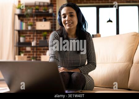 Smiling woman wearing wireless headphones and watching movie on laptop during break from remote work. Indian female freelancer seated on couch, listening to music while searching for online gigs. Stock Photo