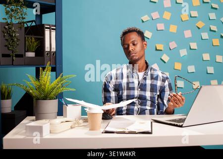 Male consultant sitting with papers in his hand, feeling overwhelmed by amount of business project workload on his desk. Stressed finance advisor holding his glasses and looking at documents. Stock Photo