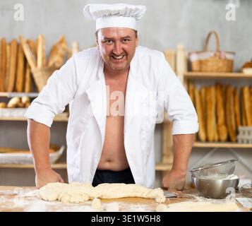 Male baker in unbuttoned uniform prepares dough, kneads it with his hands Stock Photo