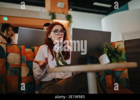 Business workers discussing a project at a laptop in a modern coworking cafe with expressive gestures Stock Photo