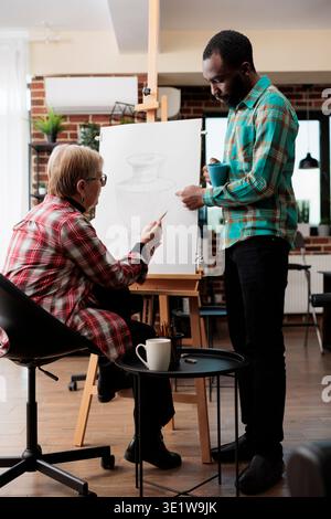 African American guy art instructor teaching senior woman to draw in class, standing near easel and explaining sketching techniques. Teacher assisting mature student during drawing lesson Stock Photo