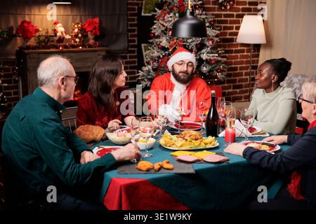Man in santa claus costume sitting at table with diverse people, enjoying festive dinner on christmas eve festivity. Young person in suit spreading positivity during december holidays. Stock Photo