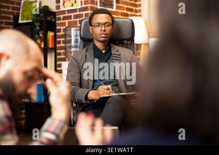 Black male counselor holds pen and observes caucasian couple during serious discussion. Professional therapy session in brick wall office, psychologist mediating relationship issues between spouses. Stock Photo