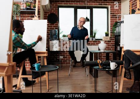 Senior man drawing teacher talking with adult students during group art class. Diverse people sitting at easels engaged in artmaking, learning to draw with tutor, making time for creative hobby Stock Photo
