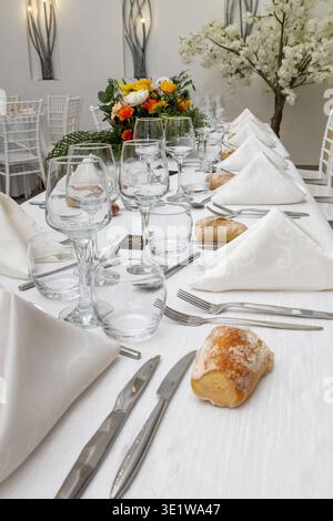Elegant wedding reception table set with refined glassware, cutlery, folded napkins, and bread rolls, complemented by a floral centerpiece. Stylish ba Stock Photo