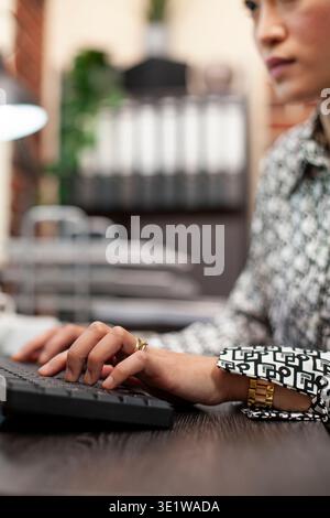 Female professional typing on computer keyboard at a modern office desk. Asian woman busy inputting project details, touching keys to update company financial information on the desktop pc. Stock Photo