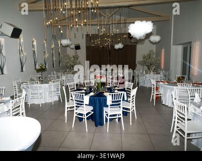 Wide view of an elegant wedding reception hall with decorated tables, white chairs, and hanging lights. Spacious modern venue arranged Stock Photo