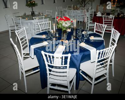 Round wedding reception table with a blue tablecloth, elegantly set with glassware, cutlery, and a floral centerpiece. Stylish indoor venue featuring Stock Photo