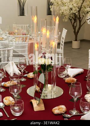 Elegant wedding reception table with a tall candle centerpiece in glass holders, surrounded by refined place settings and a red tablecloth. Stock Photo