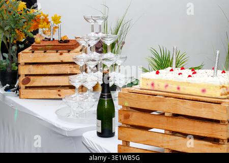 Champagne tower with stacked coupe glasses beside a dessert buffet and cake display, featuring a bottle of sparkling wine and rustic wooden decor. Stock Photo