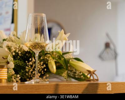Close-up of a glass of white wine placed on a table beside an elegant floral arrangement with white flowers. Stock Photo