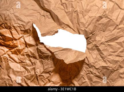 Ripped brown kraft paper forms irregular hole across crumpled textured surface. Jagged torn opening reveals empty white space, creating abstract grung Stock Photo