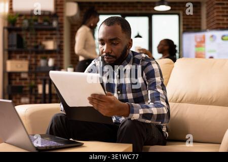 Male analyst checks printed notes and monitors company research data on laptop during marketing strategy planning. Businessman studies corporate documents on clipboard, reviewing financial charts. Stock Photo