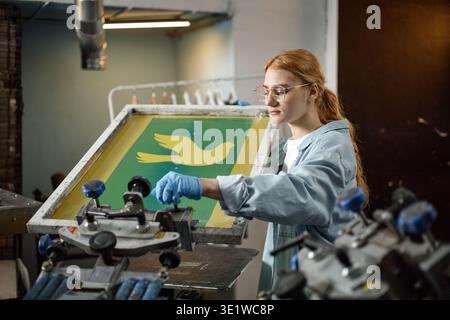 Young Adult Woman Operating Screen Printing Machine in Workshop Stock Photo
